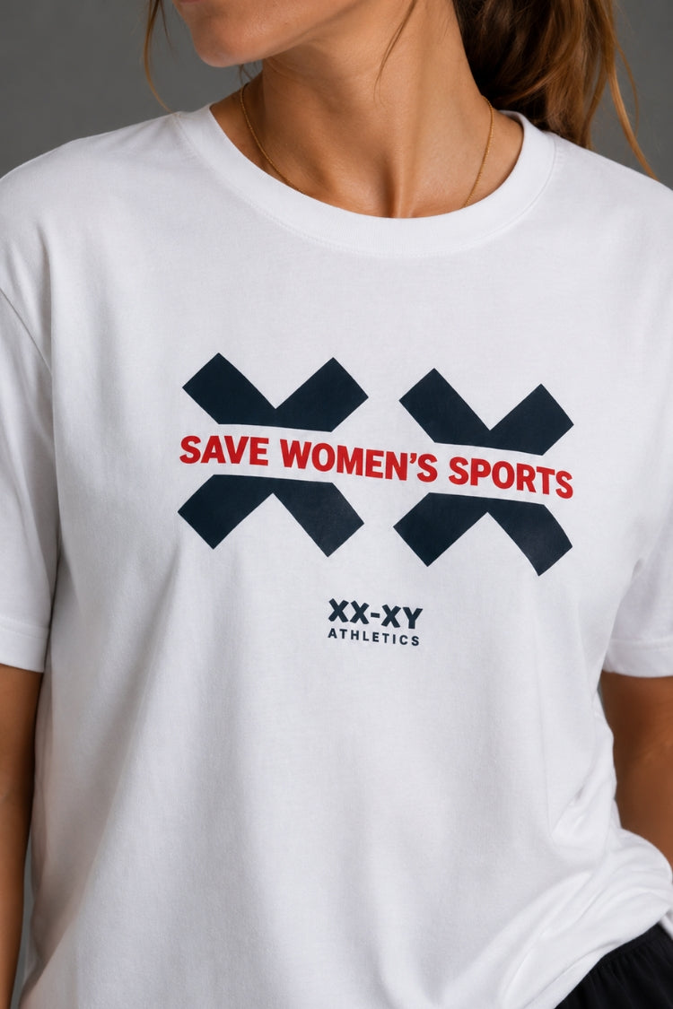 A close-up of a white crewneck t-shirt worn by a model. The shirt features a graphic with two dark navy blue 'X' logos, red text reading 'SAVE WOMEN'S SPORTS' across the center, and 'XX-XY ATHLETICS' printed in dark navy below. || White