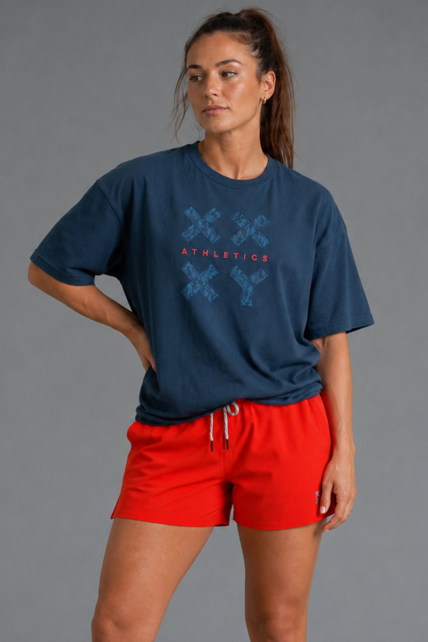 A woman modeling a navy blue athletic t-shirt with a blue and red 'ATHLETICS' graphic, paired with bright red drawstring shorts, against a solid gray studio background. || Navy