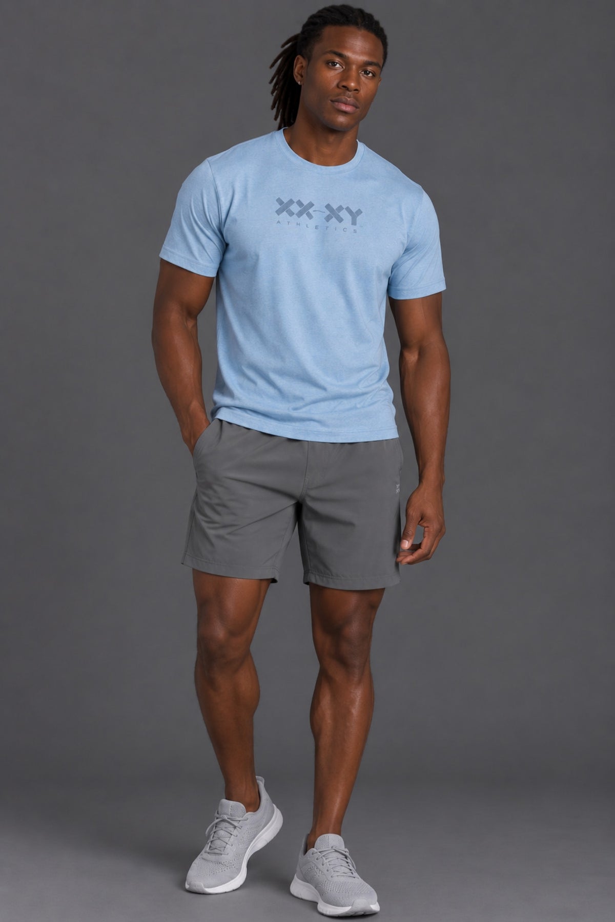 A man modeling a light blue crewneck t-shirt with a dark gray 'XX-XY' graphic on the chest, paired with gray athletic shorts and light gray sneakers, against a solid gray studio background. || Cerulean Blue Heather