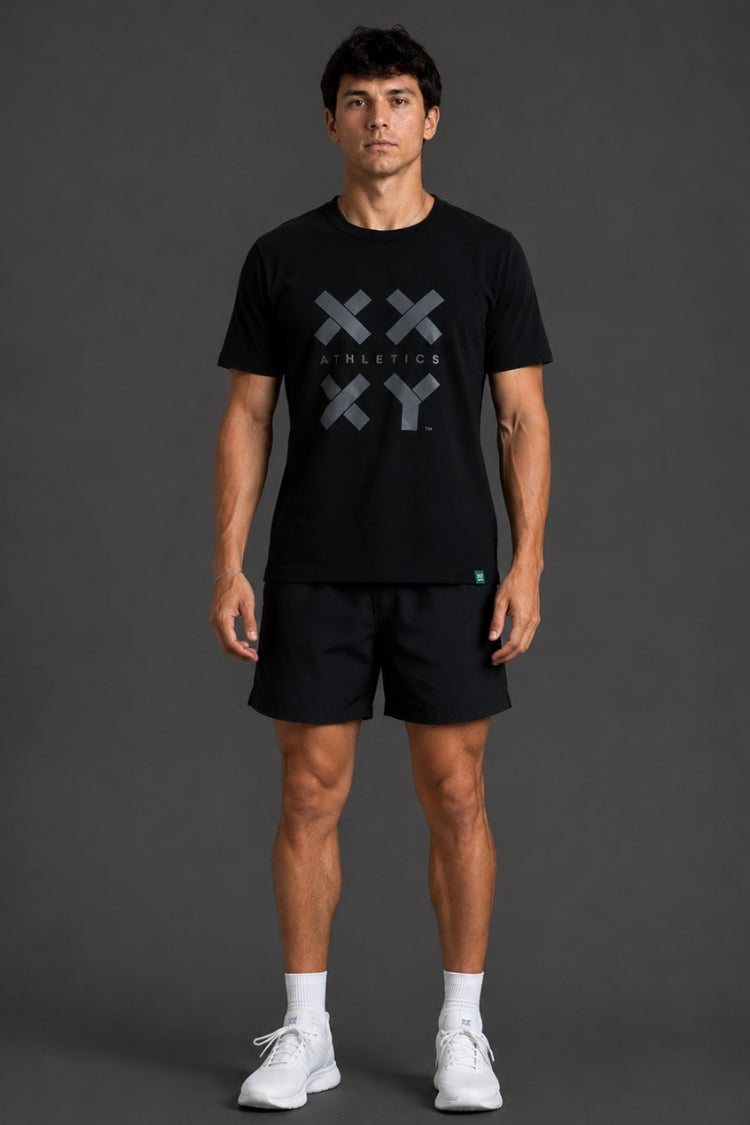 A man modeling a black short-sleeve athletic t-shirt with a gray XXY Athletics logo on the chest, paired with black shorts, white socks, and white sneakers, set against a dark gray studio background.