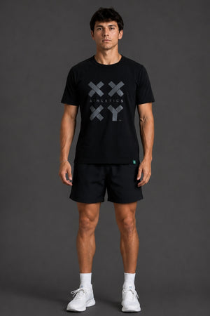 A man modeling a black short-sleeve athletic t-shirt with a gray XXY Athletics logo on the chest, paired with black shorts, white socks, and white sneakers, set against a dark gray studio background. || Black Tonal
