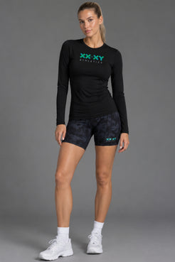 A woman modeling a black long-sleeve athletic shirt and dark patterned shorts with teal XX-XY Athletics logos, standing against a gray studio background. || Black