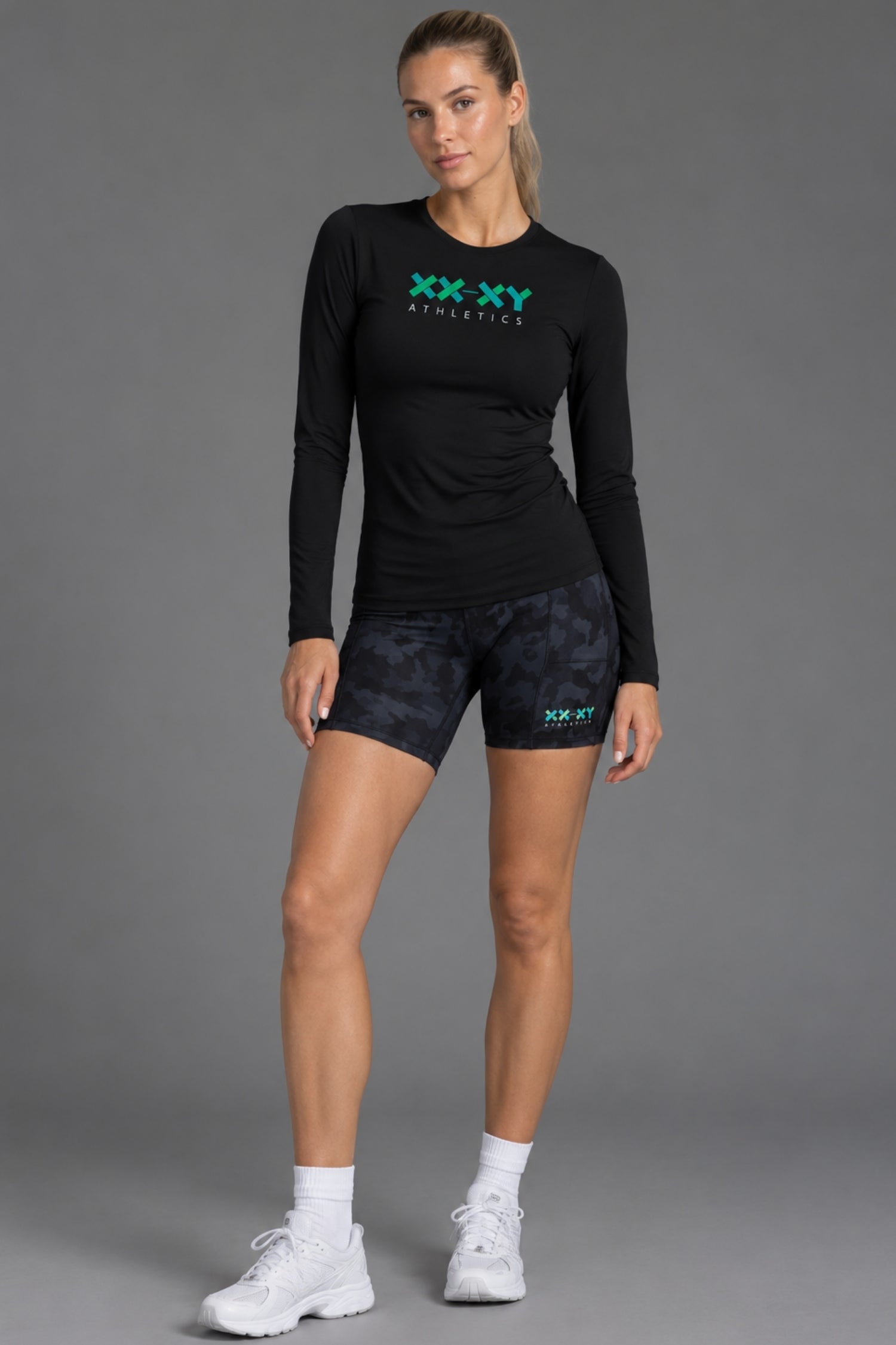 A woman modeling a black long-sleeve athletic shirt and dark patterned shorts with teal XX-XY Athletics logos, standing against a gray studio background. || Black