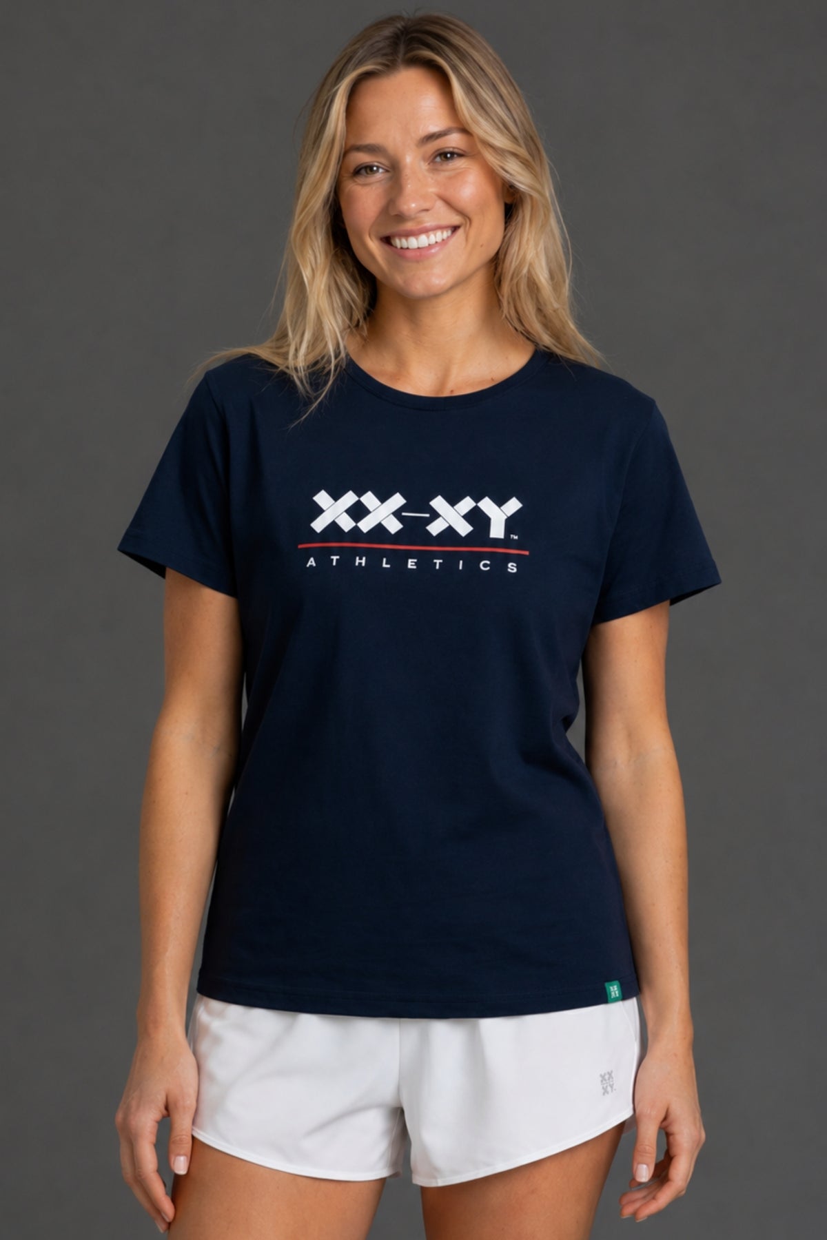A smiling blonde woman wearing a navy blue short-sleeve t-shirt with a white 'XX-X-Y ATHLETICS' logo on the chest, paired with white athletic shorts, set against a solid dark gray studio background.
