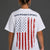 A woman viewed from the back wearing a white graphic T-shirt with the text 'SAVE WOMEN'S SPORTS' above a distressed American flag design in red, white, and blue. She is also wearing black shorts against a dark gray studio background. || White