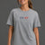 A woman wearing a heather gray short-sleeve t-shirt with a 'XX-XY ATHLETICS' logo printed on the chest, paired with black bike shorts, against a solid dark gray studio background. || Heather Gray