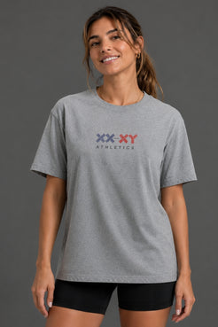 A woman wearing a heather gray short-sleeve t-shirt with a 'XX-XY ATHLETICS' logo printed on the chest, paired with black bike shorts, against a solid dark gray studio background. || Heather Gray