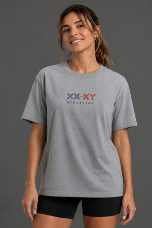A woman wearing a heather gray short-sleeve t-shirt with a 'XX-XY ATHLETICS' logo printed on the chest, paired with black bike shorts, against a solid dark gray studio background. || Heather Gray