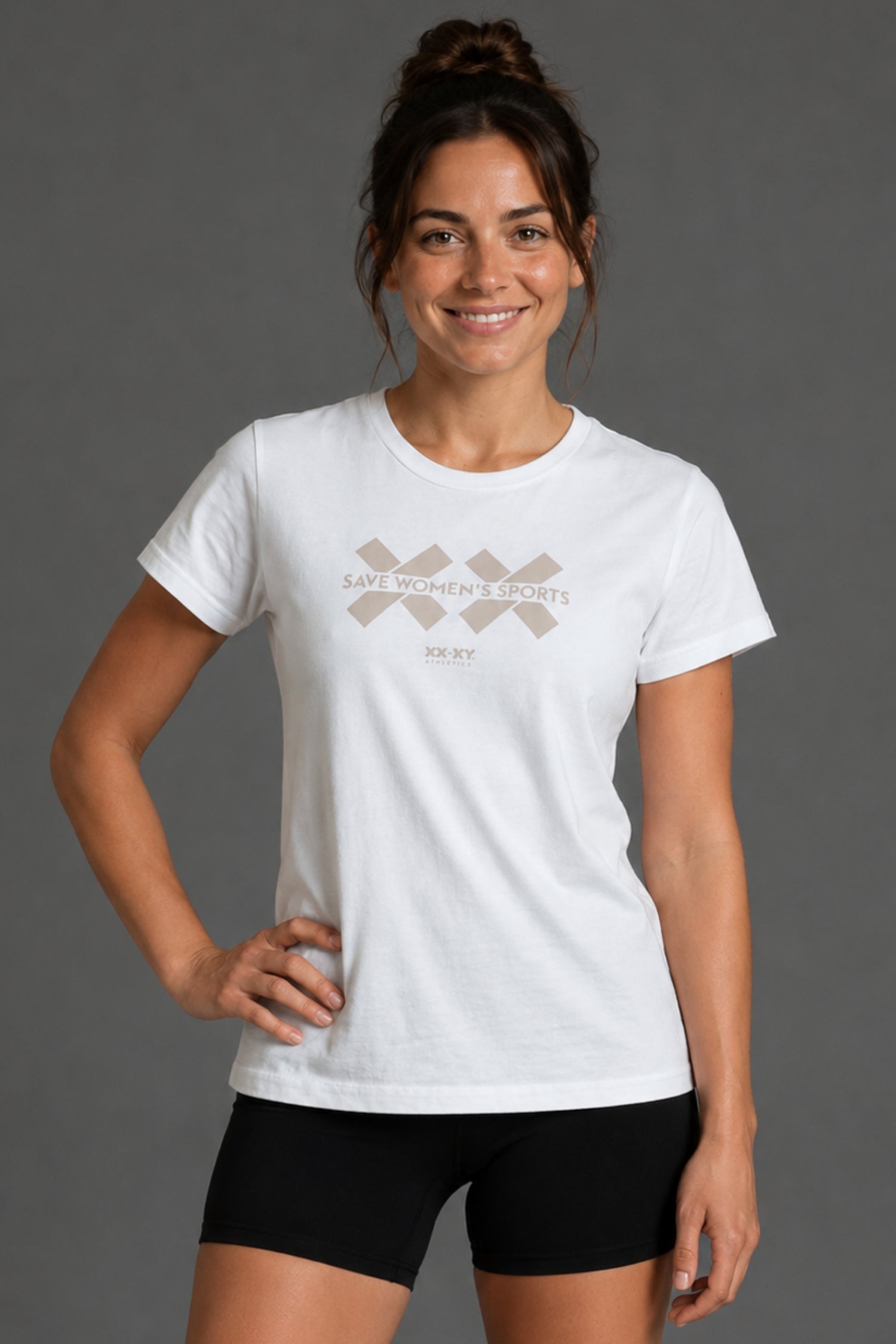 A woman modeling a white short-sleeve crewneck t-shirt with a gray 'Save Women's Sports NEXT' logo on the chest, paired with black athletic shorts, against a solid gray studio background. || Taupe