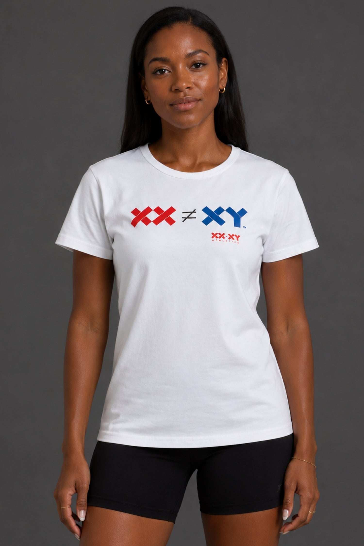 A woman modeling a white short-sleeve crewneck t-shirt with a red and blue chest graphic reading 'XX ≠ ♦ XY' and 'XX-XY' underneath, paired with black bike shorts against a dark gray studio background.