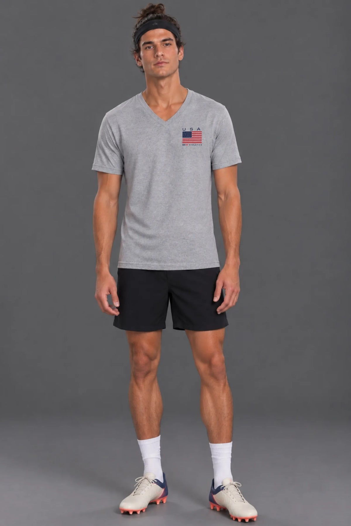 Male model standing against a gray studio background wearing the heather gray XX-XY Athletics USA Flag V-Neck Tee, black athletic shorts, white socks, and soccer cleats. || Light Heather Gray