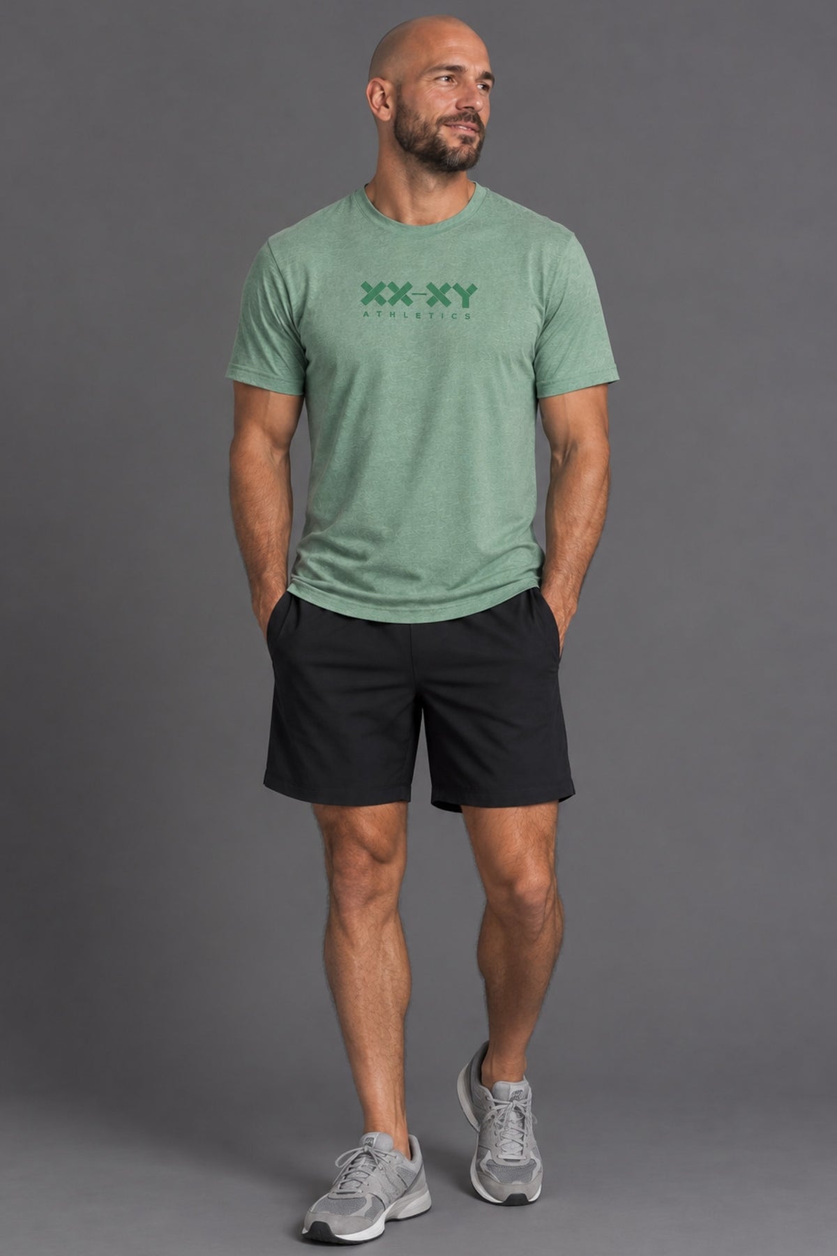 A man modeling a light green heathered t-shirt with a dark green 'XX XY' graphic on the chest, paired with black athletic shorts and gray sneakers, against a solid gray studio background.