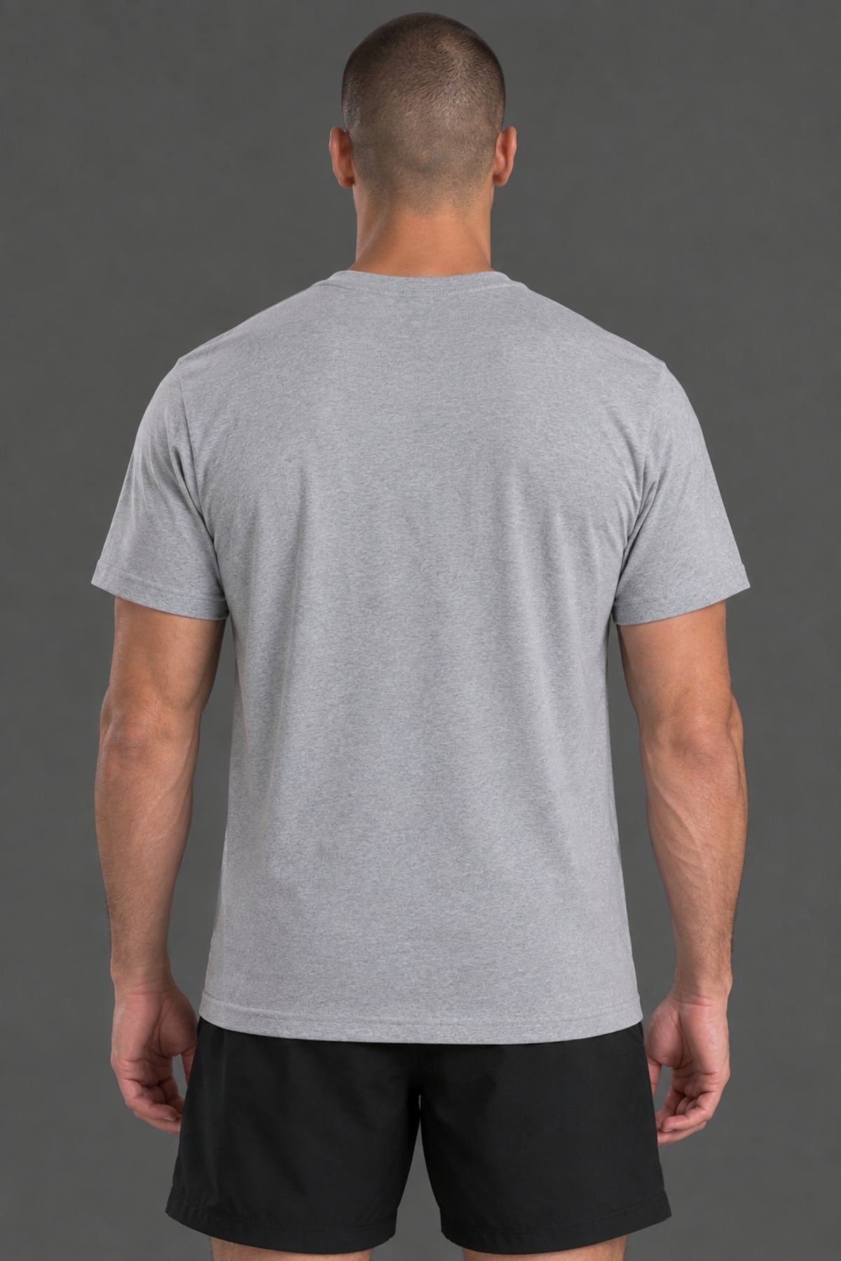 A man viewed from behind wearing a light gray heather crewneck t-shirt and black athletic shorts, standing against a solid dark gray studio background. || Light Heather Gray