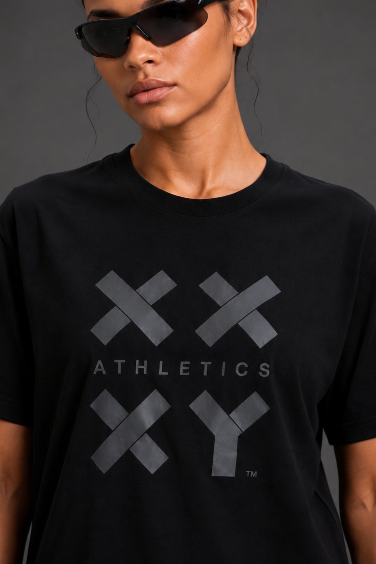 A close-up of a model wearing a black crewneck t-shirt with a dark gray graphic on the front featuring the text 'ATHLETICS' and geometric 'XXY' shapes. The model is also wearing black sunglasses. || Black Tonal