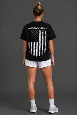 A woman viewed from the back wearing a black short-sleeve T-shirt with a distressed American flag graphic and the words 'SAVE WOMEN'S SPORTS' printed across the upper back, paired with white athletic shorts, socks, and sneakers against a dark gray studio background. || Black