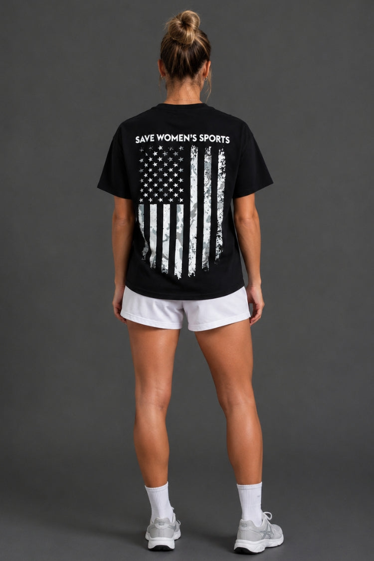 A woman viewed from the back wearing a black short-sleeve T-shirt with a distressed American flag graphic and the words 'SAVE WOMEN'S SPORTS' printed across the upper back, paired with white athletic shorts, socks, and sneakers against a dark gray studio background. || Black