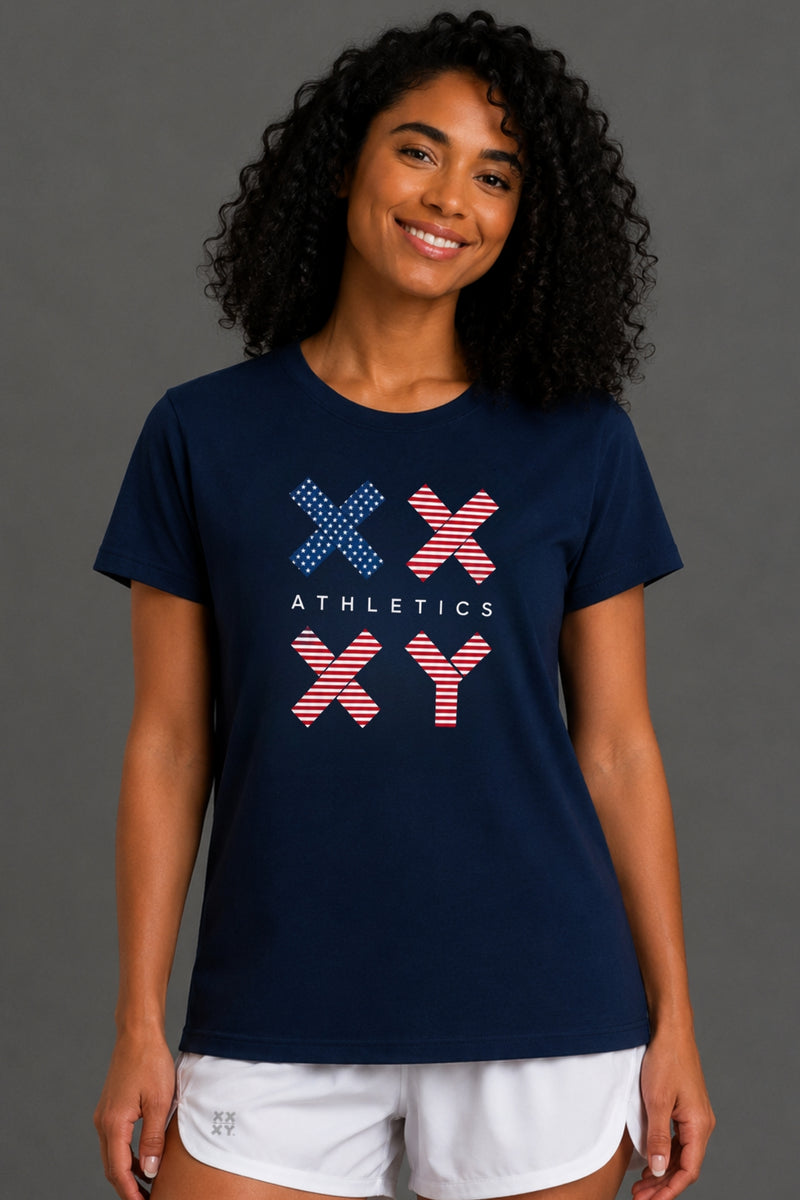 A woman with curly black hair wearing a navy blue short-sleeve t-shirt with a red, white, and blue graphic on the front, paired with white shorts, against a gray studio background. || Dark Navy