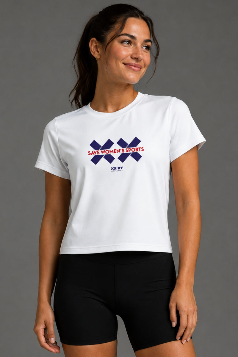 A woman wearing a white cropped t-shirt with a 'SAVE WOMEN'S SPORTS' graphic in red and navy blue, paired with black athletic shorts, against a solid gray studio background. || White