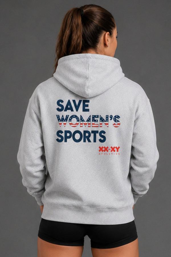 A woman viewed from behind wearing a light gray hoodie with a graphic on the back that reads 'SAVE WOMEN'S SPORTS' in navy and red, plus 'XX-XY SPORTS' below. She is also wearing black athletic shorts against a plain gray studio background. || Heather Gray
