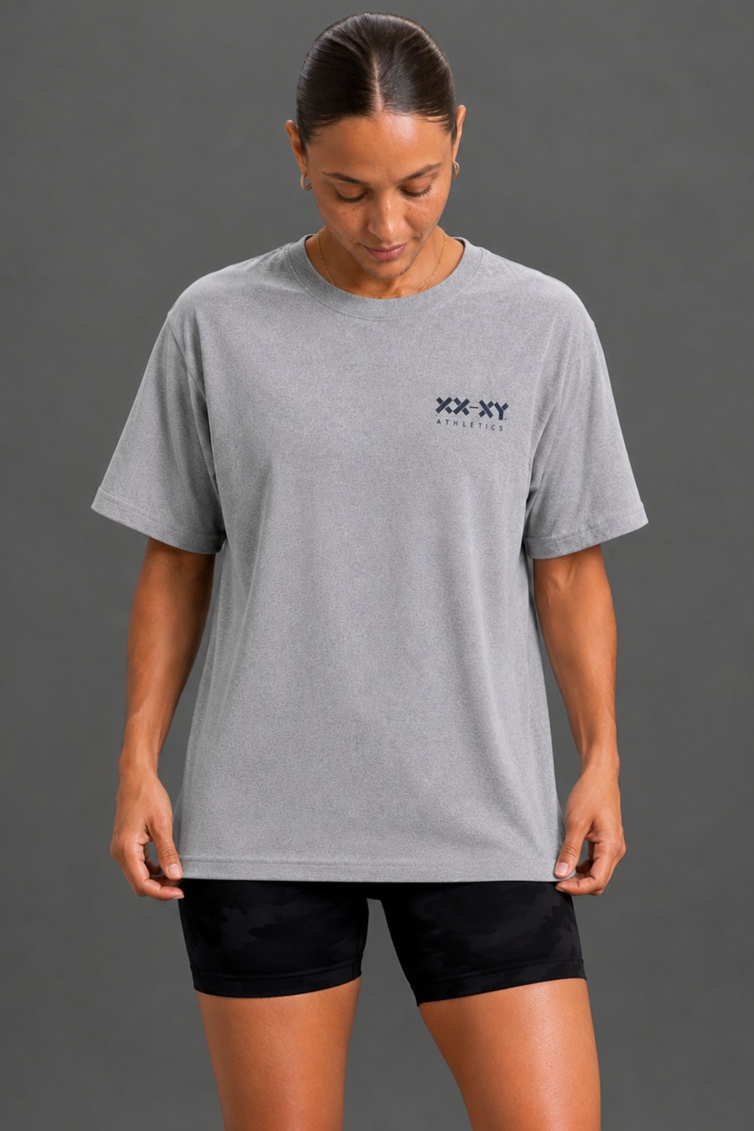 A woman wearing a light gray heather crewneck t-shirt with a small black 'XX XY ATHLETICS' logo on the left chest, paired with black athletic shorts, against a solid dark gray studio background. || Heather Gray