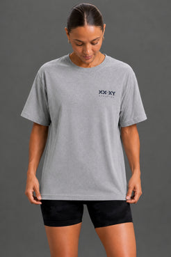 A woman wearing a light gray heather crewneck t-shirt with a small black 'XX XY ATHLETICS' logo on the left chest, paired with black athletic shorts, against a solid dark gray studio background. || Heather Gray