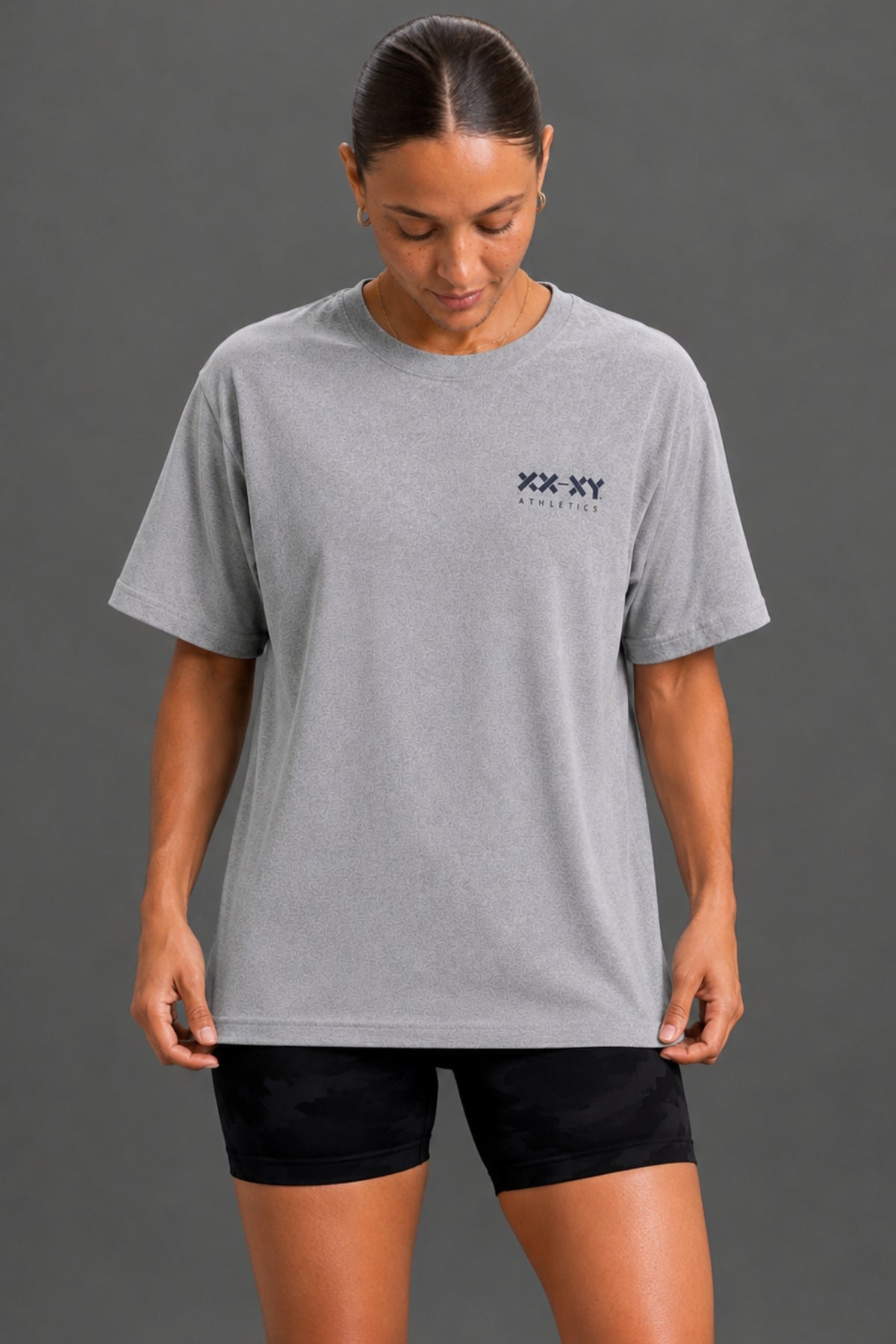 A woman wearing a light gray heather crewneck t-shirt with a small black 'XX XY ATHLETICS' logo on the left chest, paired with black athletic shorts, against a solid dark gray studio background. || Heather Gray