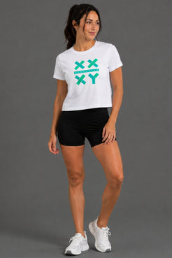 A woman modeling a white cropped t-shirt with a teal 'XX XY' graphic on the front, paired with black athletic shorts and white sneakers, against a solid gray studio background. || White