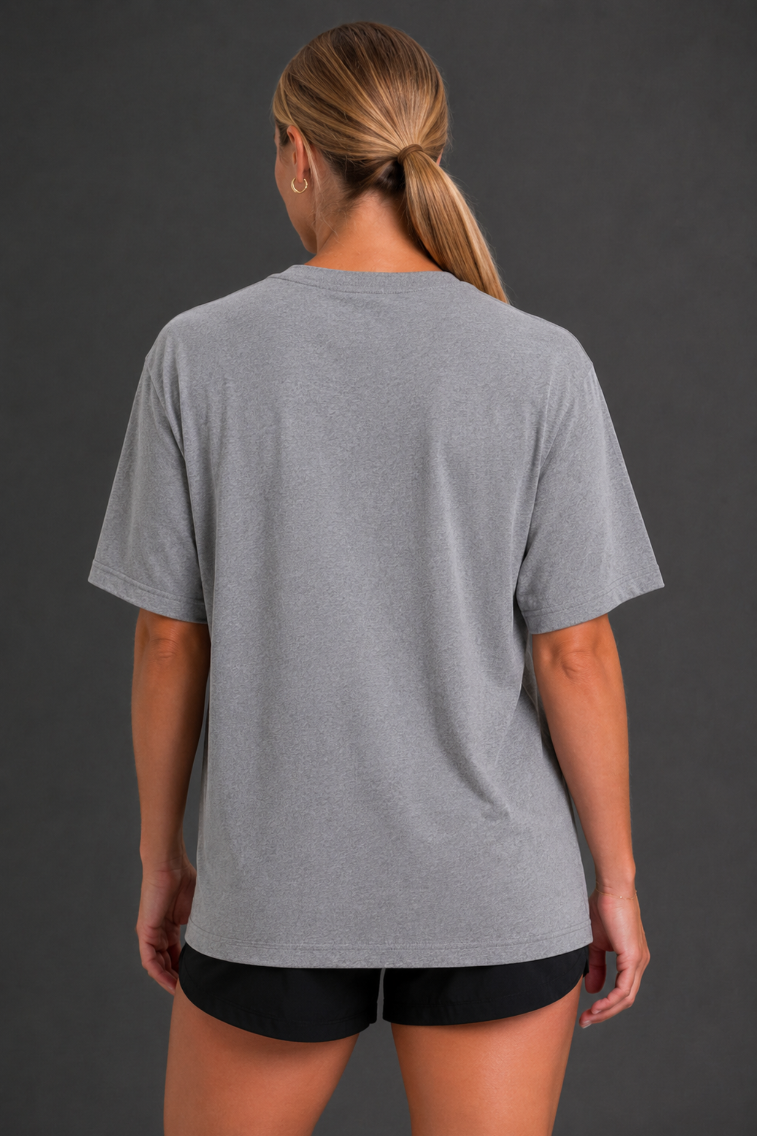 A woman viewed from the back wearing a heather gray short-sleeve t-shirt and black athletic shorts against a dark gray studio background.