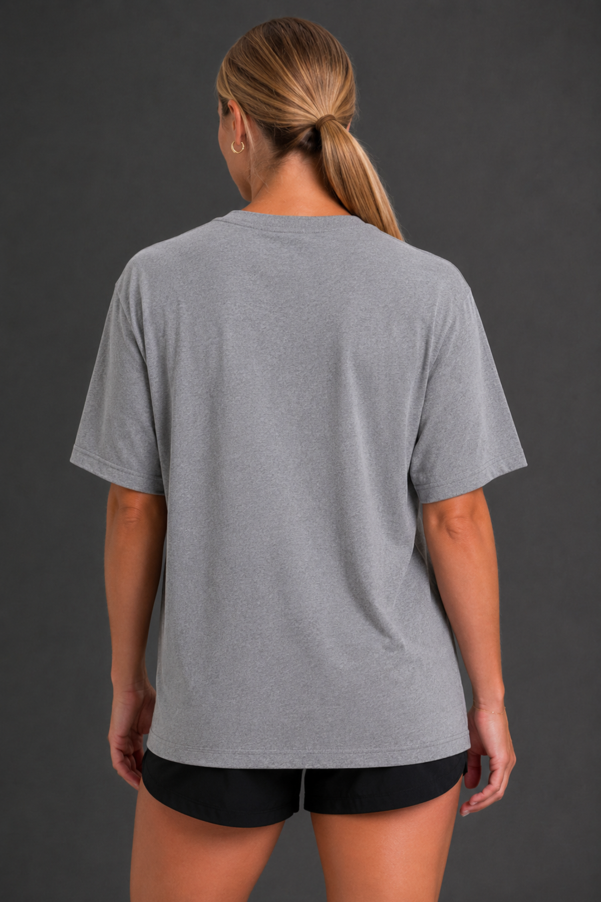 A woman viewed from the back wearing a heather gray short-sleeve t-shirt and black athletic shorts against a dark gray studio background. || Heather Gray