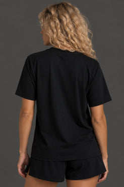 A woman with curly blonde hair is shown from behind wearing a black short-sleeve t-shirt and matching black shorts against a dark gray studio background. || Black