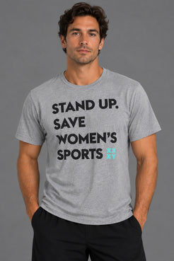 A man wearing a heather gray graphic t-shirt with the text 'STAND UP. SAVE WOMEN'S SPORTS' in black, with a small light blue logo accent, against a solid gray studio background. || Heather Gray