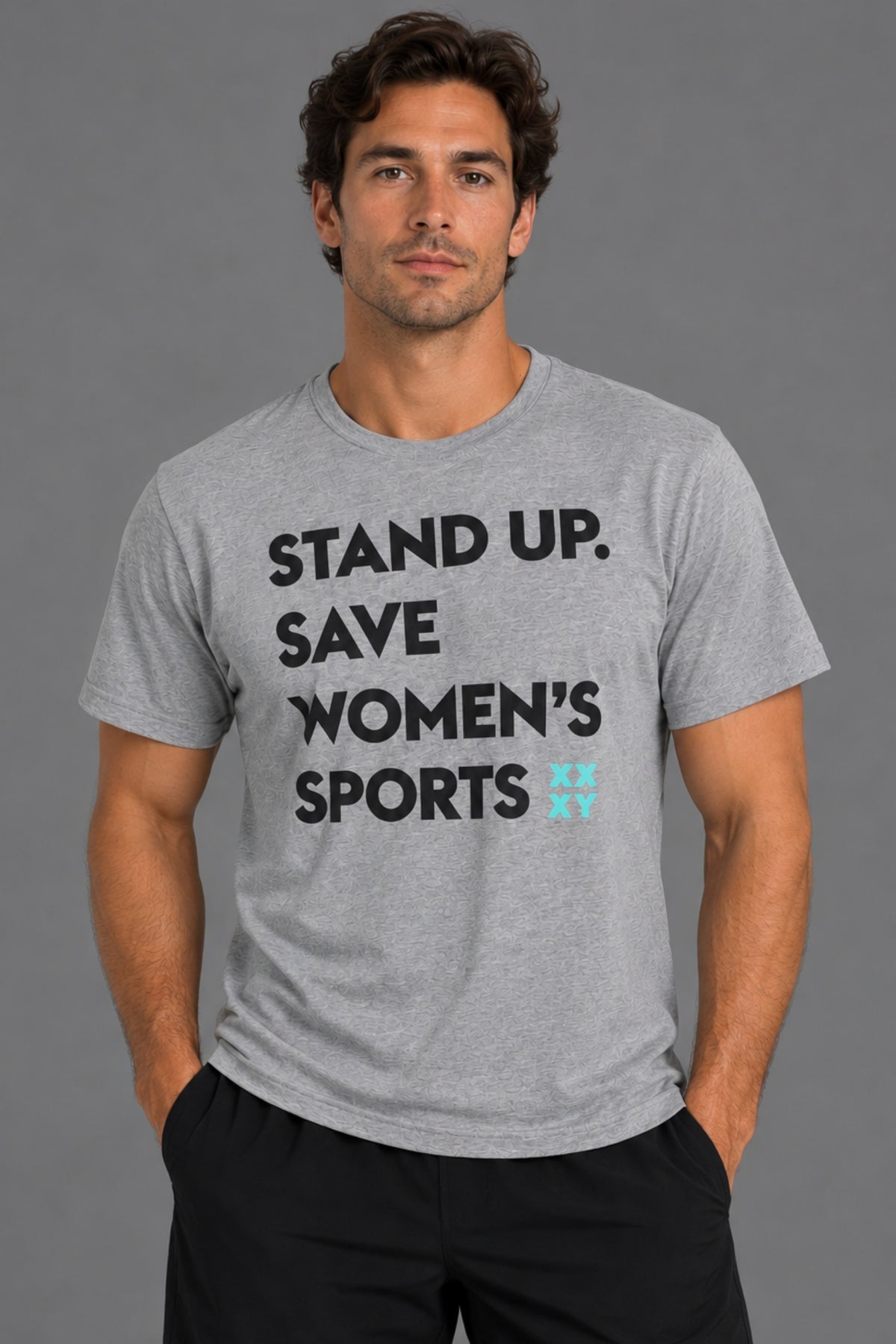 A man wearing a heather gray graphic t-shirt with the text 'STAND UP. SAVE WOMEN'S SPORTS' in black, with a small light blue logo accent, against a solid gray studio background. || Heather Gray