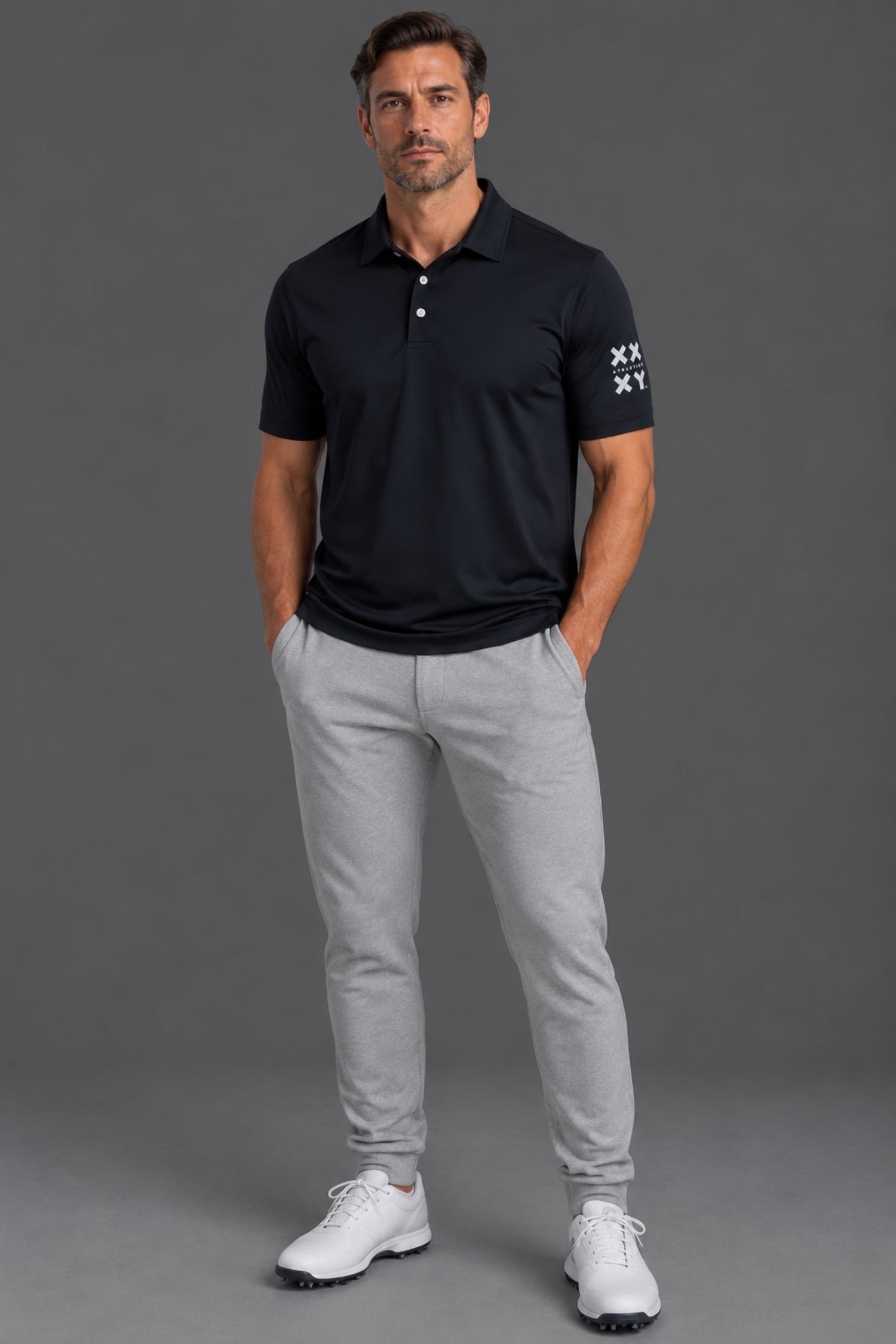 A man modeling a dark navy polo shirt with white embroidered logos on the sleeve, paired with light gray jogger pants and white golf shoes, against a solid gray studio background.