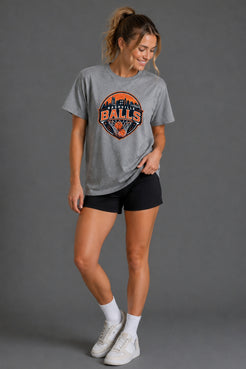 A woman modeling a heather gray graphic t-shirt with an orange and blue 'Houston Rockets' logo, paired with black athletic shorts, white socks, and white sneakers against a solid gray studio background. || Heather Gray