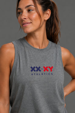 A woman wearing a heather gray sleeveless muscle tank top with a 'XX-XY ATHLETICS' logo printed on the chest in navy and red, set against a solid dark gray studio background. || Dark Heather Gray