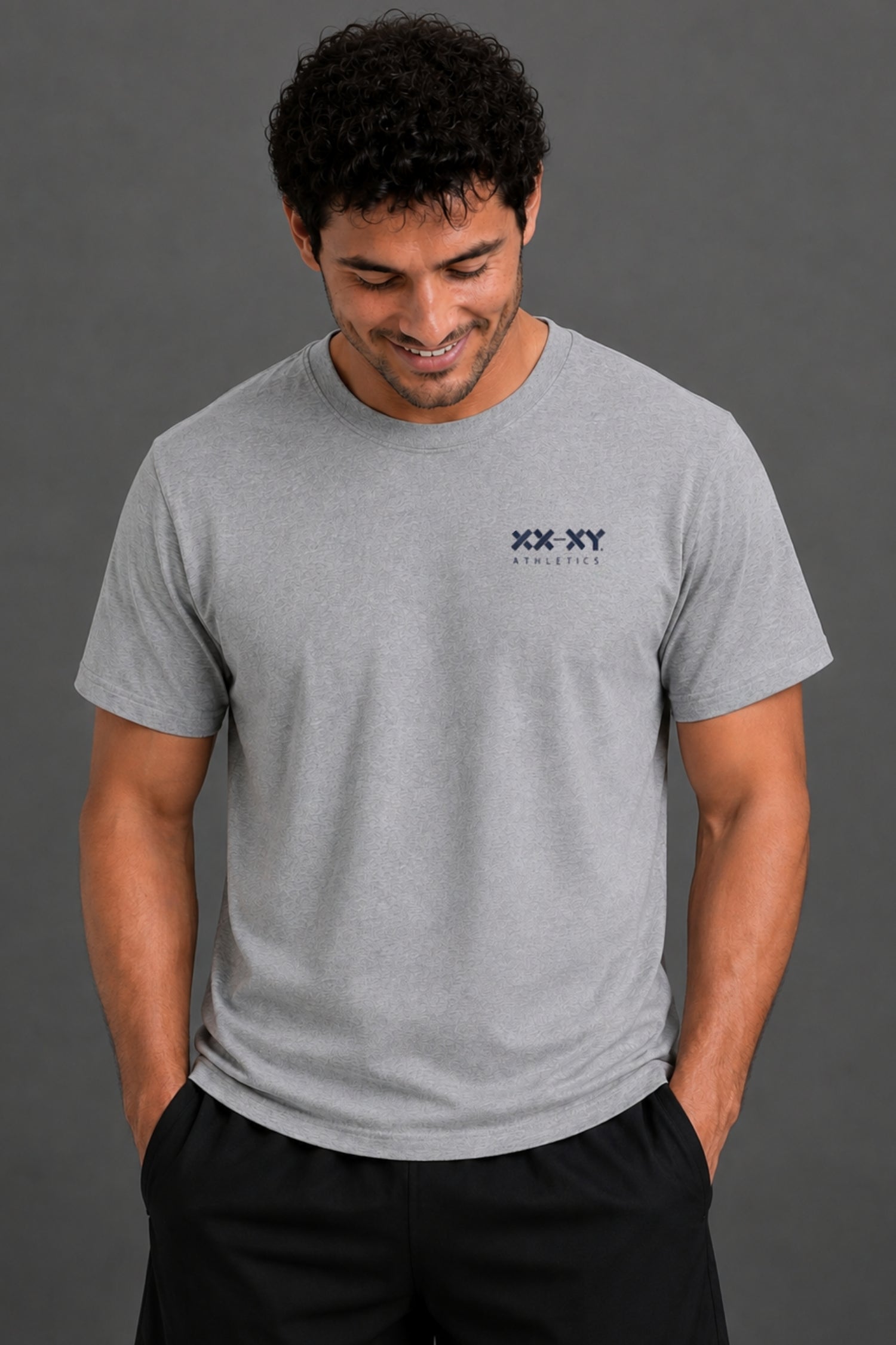 A man wearing a light gray crewneck t-shirt with a small black 'XX-XY' logo on the left chest, paired with black pants, set against a solid dark gray background.