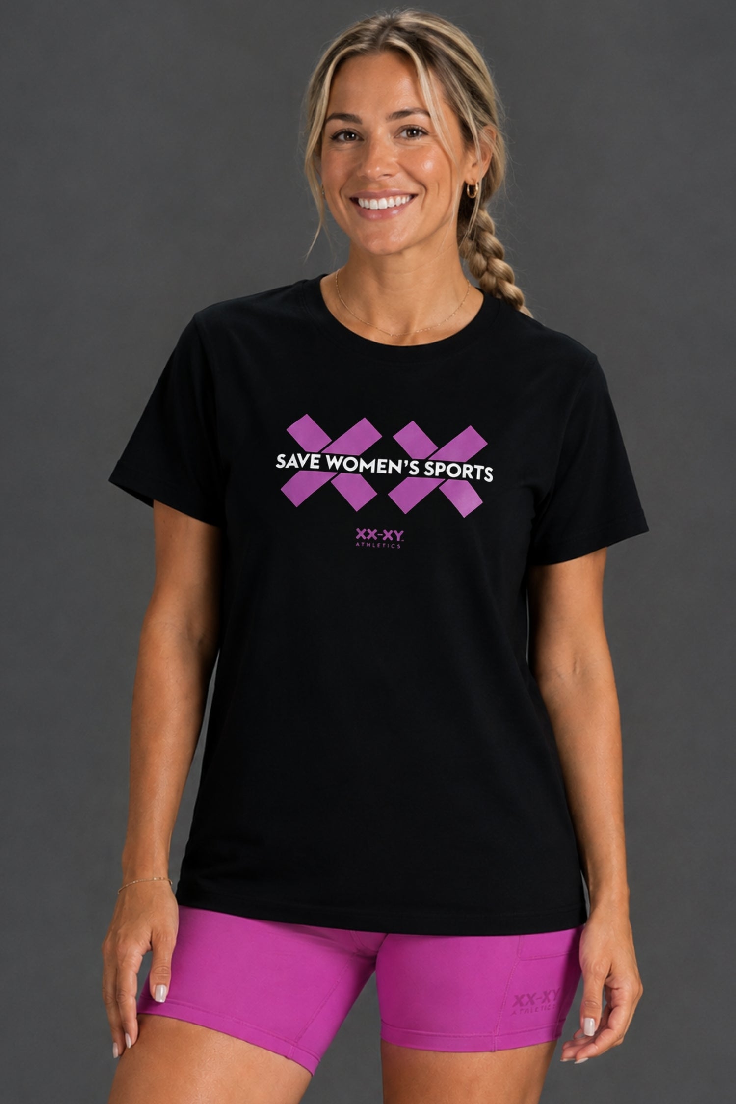 A woman modeling a black short-sleeve t-shirt with a purple graphic that reads 'SAVE WOMEN'S SPORTS' and 'XX.XX' below it, paired with bright pink athletic shorts against a dark gray studio background. || Bright Fuchsia