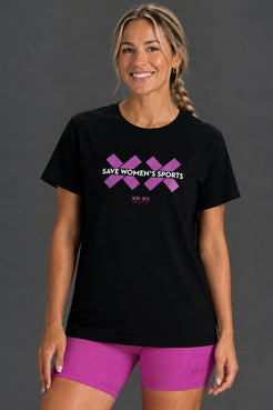 A woman modeling a black short-sleeve t-shirt with a purple graphic that reads 'SAVE WOMEN'S SPORTS' and 'XX.XX' below it, paired with bright pink athletic shorts against a dark gray studio background. || Bright Fuchsia