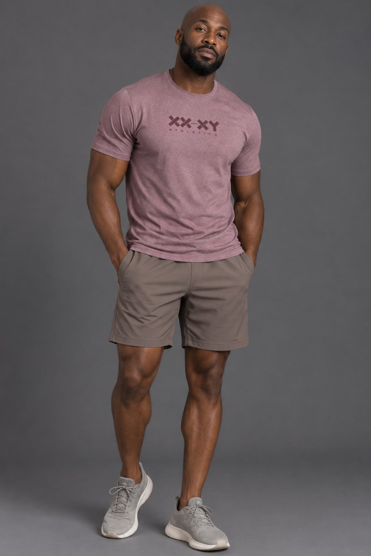 A muscular man modeling a mauve-pink short-sleeve t-shirt with a dark 'XX-XY' graphic on the chest, paired with taupe-brown athletic shorts and gray sneakers, against a solid gray studio background. || Burgundy Heather