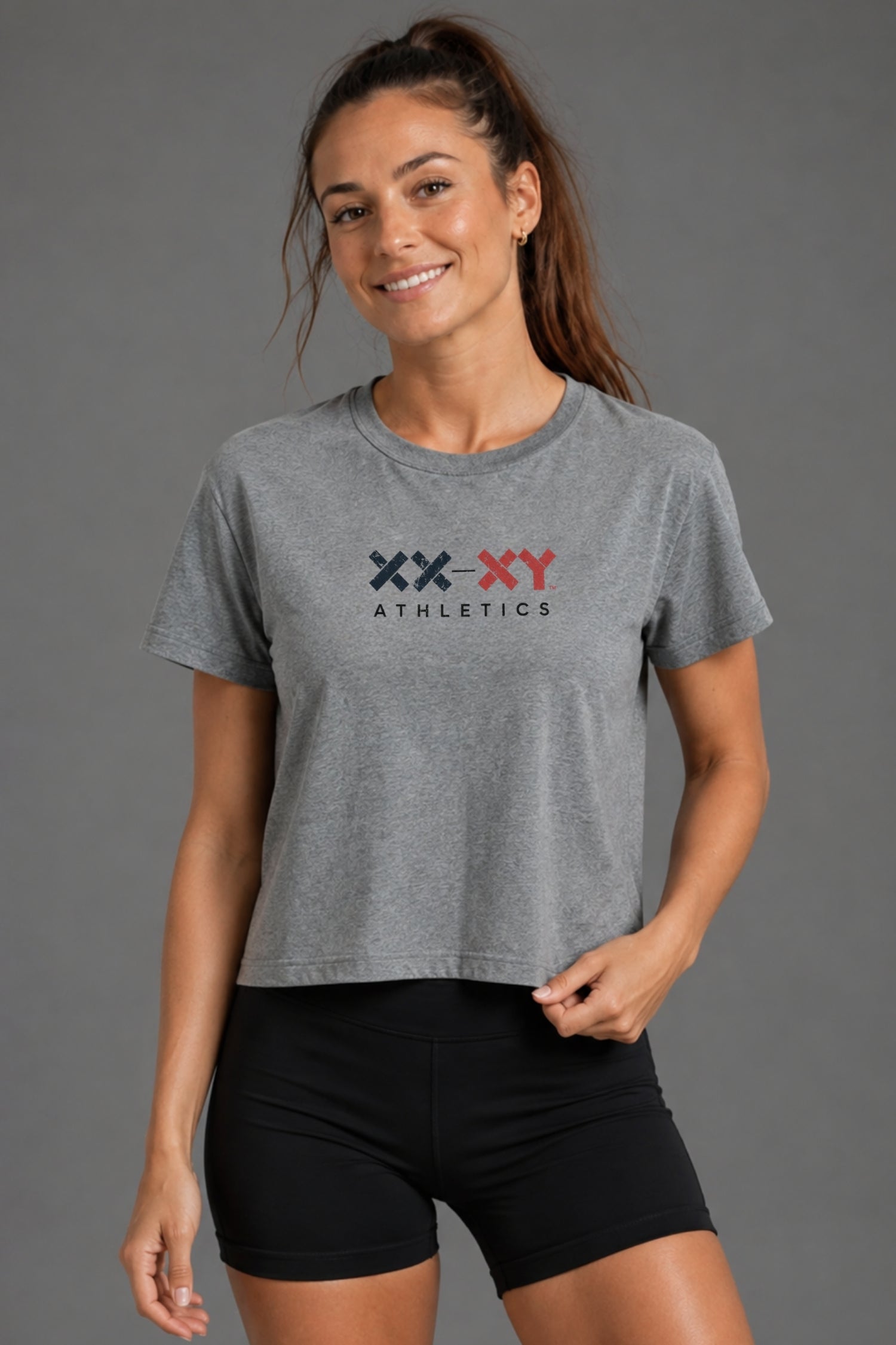A woman wearing a heather gray cropped t-shirt with a navy and red 'XX-XY ATHLETICS' logo on the chest, paired with black athletic shorts, against a solid gray studio background.