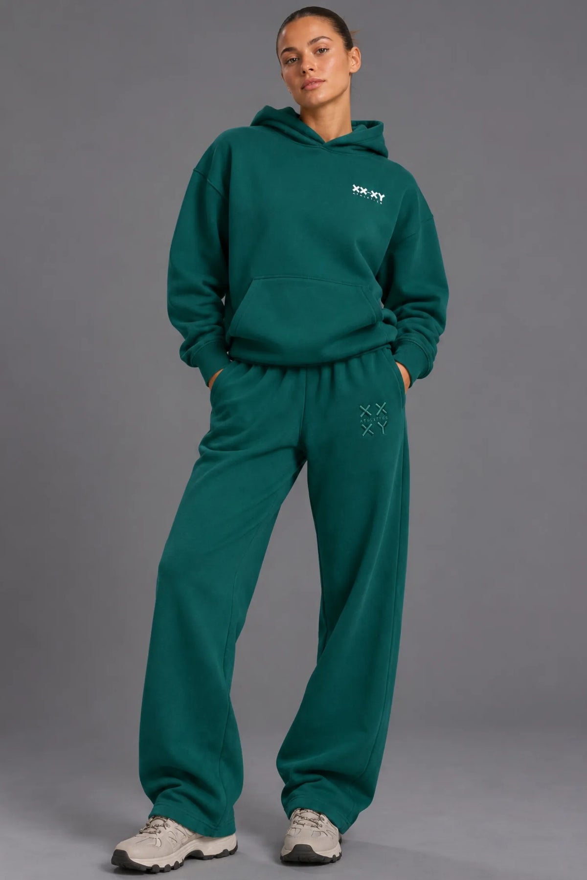 Woman wearing a teal XX-XY Athletics hoodie and matching sweatpants against a gray studio background. || Pine