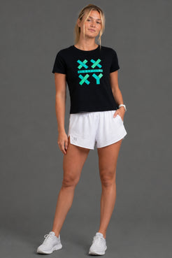 A woman modeling a black short-sleeve t-shirt with a teal 'XX XY' graphic on the chest, paired with white athletic shorts and white sneakers, against a solid gray studio background. || Black