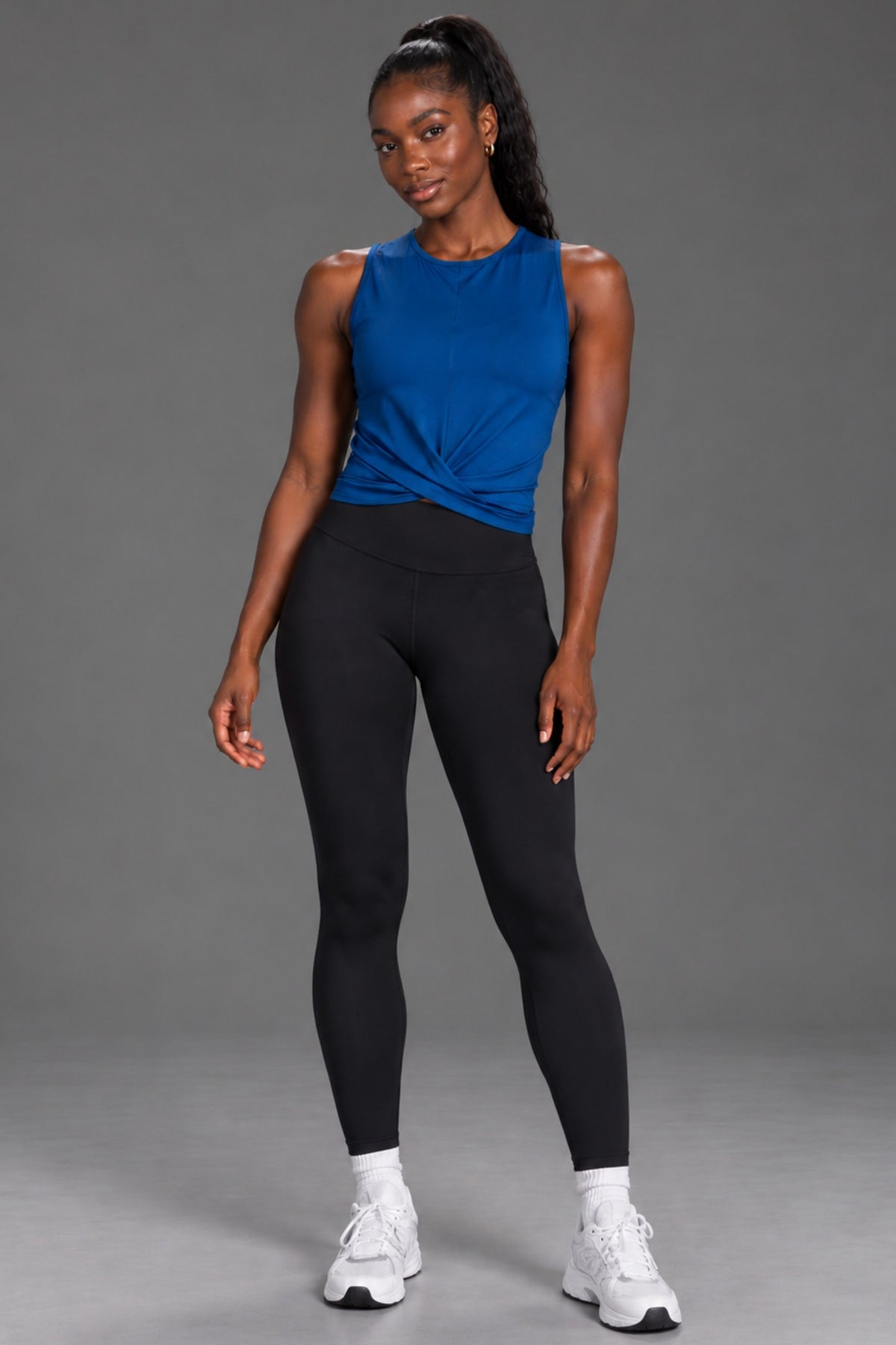 Black female model wearing a blue twist-front tank and black XX-XY Athletics leggings on a gray studio background.