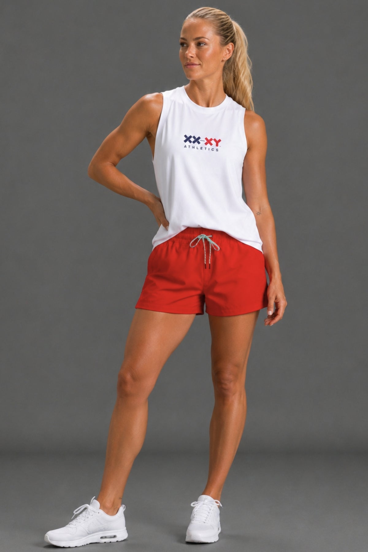 A woman modeling a white sleeveless athletic tank top with a red and blue 'XX XY GYMNASIUM' logo on the chest, paired with bright red athletic shorts and white sneakers, against a solid gray studio background.