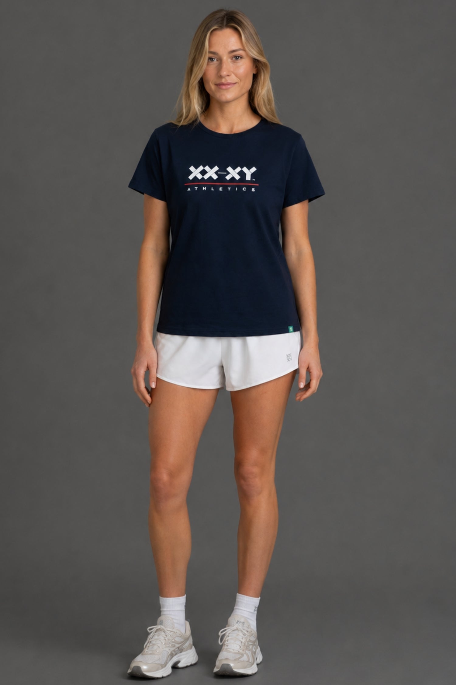 A woman modeling a navy blue short-sleeve athletic t-shirt with a white 'XX-XY ATHLETICS' logo on the chest, paired with white athletic shorts, white socks, and white sneakers, against a solid gray studio background.
