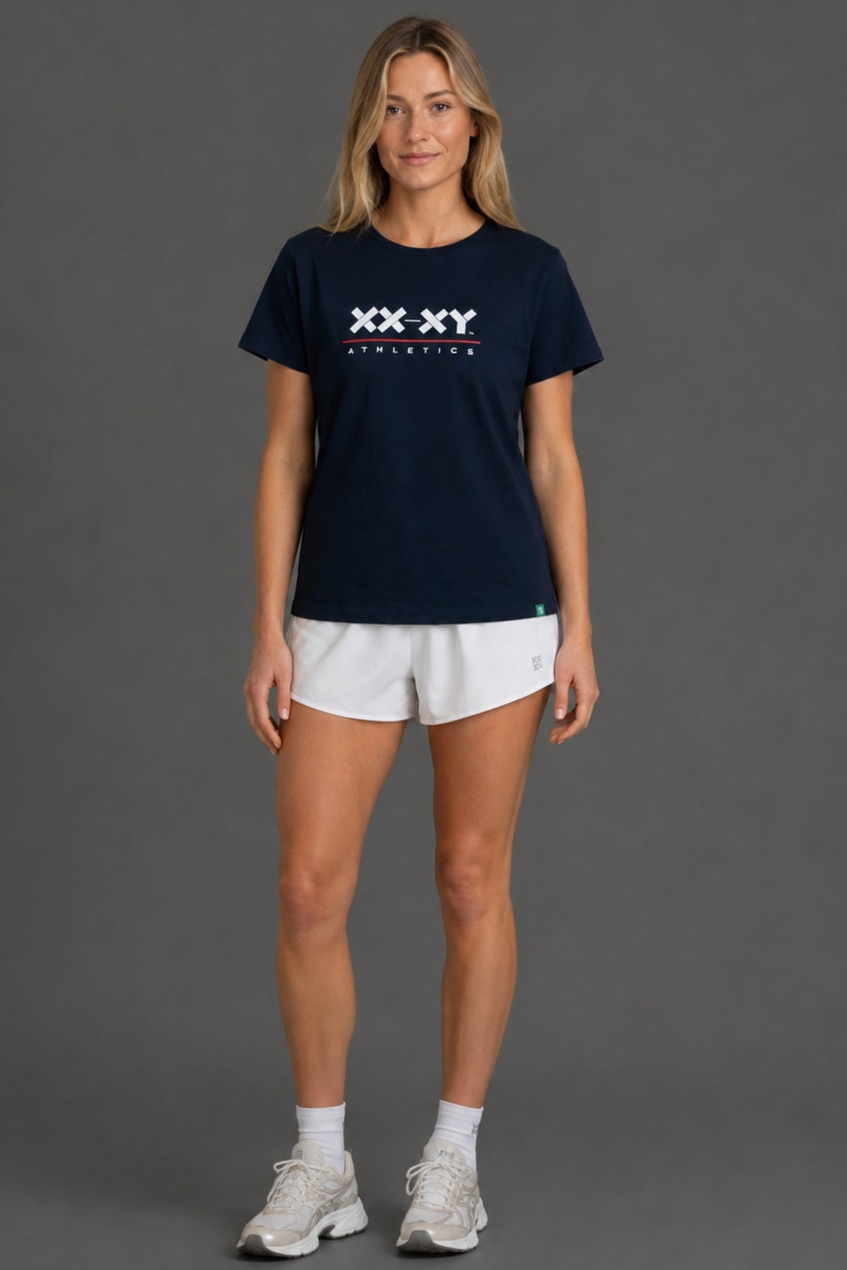 A woman modeling a navy blue short-sleeve athletic t-shirt with a white 'XX-XY ATHLETICS' logo on the chest, paired with white athletic shorts, white socks, and white sneakers, against a solid gray studio background. || Navy
