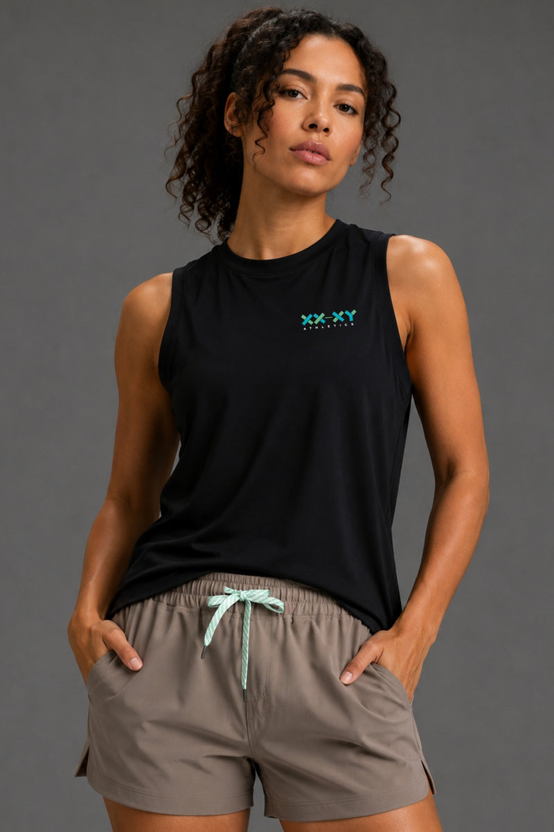 A woman modeling a black sleeveless athletic tank top with a small green XX.YY logo on the chest, paired with taupe drawstring shorts, against a solid dark gray studio background. || Black