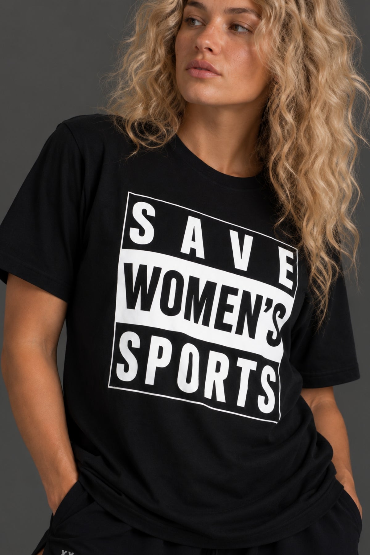 A woman wearing a black short-sleeve graphic T-shirt with the slogan 'SAVE WOMEN'S SPORTS' printed in bold white block letters on the front, photographed against a dark gray studio background.
