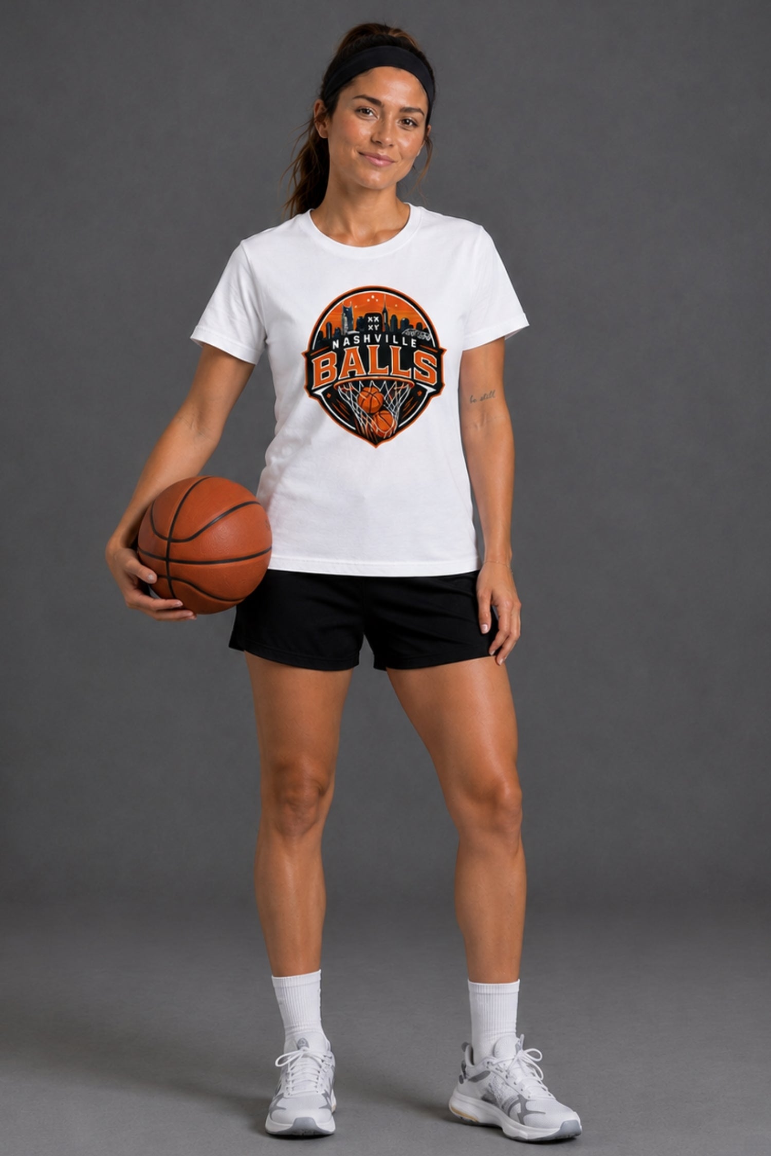 A woman modeling a white crewneck t-shirt with a circular orange and black 'BALLS' graphic, paired with black athletic shorts, white socks, and white sneakers, holding a basketball against a dark gray studio background.