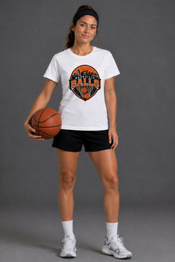 A woman modeling a white crewneck t-shirt with a circular orange and black 'BALLS' graphic, paired with black athletic shorts, white socks, and white sneakers, holding a basketball against a dark gray studio background. || White